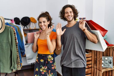 Satış mağazasında alışveriş torbaları tutan genç İspanyol çift merhaba diyor mutlu ve gülümsüyor, dostça bir hoş geldin jesti 