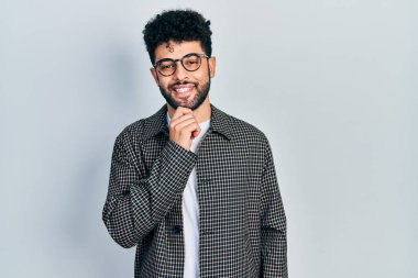 Sakallı, gözlüklü, kameraya kendinden emin, kolları çapraz ve çenesi havada gülümseyen genç bir Arap. Olumlu düşünmek. 