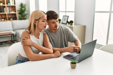 Kafkasyalı genç çift evde masada dizüstü bilgisayar kullanarak mutlu bir şekilde gülümsüyor..