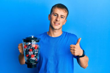 Kafkasyalı genç adam elinde meyve püresi, gülümseyen mutlu ve pozitif bir şekilde gülümsüyor, başparmağıyla mükemmel ve onay işareti yapıyor. 