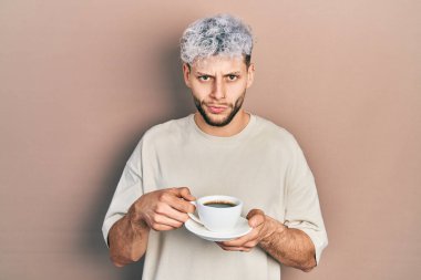 Modern boyalı saçlı genç İspanyol bir adam bir fincan kahve içiyor şüpheci ve gergin, sorun yüzünden surat asıyor. negatif kişi. 