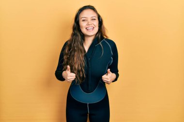 Genç İspanyol kız neoprene üniforması giyiyor. Başarı işareti el hareketleriyle olumlu bir hareket yapıyor. Gülümsüyor ve mutlu. Neşeli ifade ve kazanan jest. 