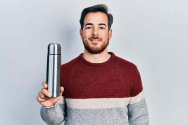 Sakallı yakışıklı beyaz adam termo tutuyor. Pozitif ve mutlu görünüyor. Kendine güvenen bir gülümsemeyle dişlerini gösteriyor. 