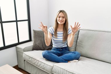 Asyalı genç bir kadın evdeki kanepede oturuyor. Sekiz numaralı parmaklarıyla işaret ediyor. Kendinden emin ve mutlu bir şekilde gülümsüyor.. 