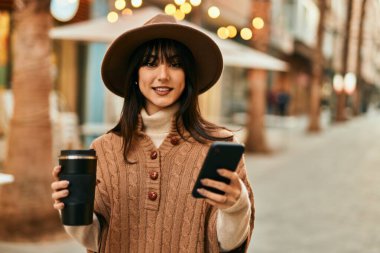 Kışlık şapkalı esmer kadın akıllı telefon kullanıyor ve şehirde bir fincan kahve içiyor.