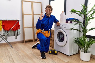 Çamaşır makinesinde çalışan Latin asıllı genç adam parmaklarıyla imza atıyor, gülümseyerek mükemmel bir sembol oluşturuyor. 