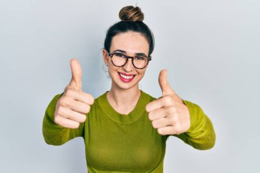 Gündelik kıyafetler giyen ve gözlük takan genç İspanyol kız elinle olumlu jest yapıyor, gülümsüyor ve başarı için mutlu. kazanan jest. 