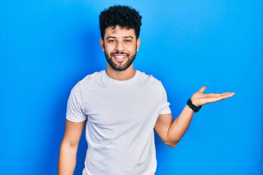 Sakallı, beyaz tişörtlü, neşeli gülümseyen, avuç içi dolu bir şekilde kameraya bakan genç bir Arap.. 