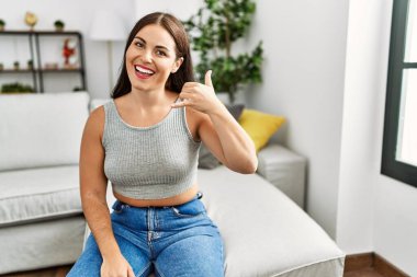 Genç esmer kadın evdeki kanepede oturmuş gülümsüyor telefonla el ve parmaklarıyla telefonla konuşuyor gibi hareket ediyor. iletişim kavramları. 