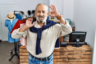 Butik dükkanında alışveriş poşetleri tutan yakışıklı bir adam gülümsüyor ve el ve parmaklarıyla 