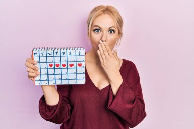 Elinde kalp takvimi olan genç sarışın bir kadın. Ağzı eliyle kapatıyor. Şok olmuş ve hata yapmaktan korkuyor. şaşırmış ifade 