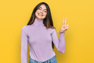 Balıkçı yaka kazak giyen genç ve güzel bir kız kendini beğenmiş ve mutlu bir şekilde gülümserken parmaklarıyla iki numarayı gösteriyor.. 