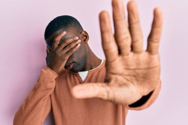Genç, Afro-Amerikan bir adam, günlük kıyafetler giyiyor, elleriyle gözleri örtüyor ve üzgün ve korku ifadeleriyle jestleri durduruyor. utangaç ve olumsuz kavram. 