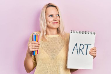 Güzel, beyaz, sarışın bir kadın sanat defteri ve renkli kalemler tutuyor. Gülümsüyor, yan tarafa bakıyor ve düşünüyor.. 