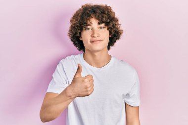 Yakışıklı genç adam, günlük beyaz tişört giyip mutlu mesut el hareketi yapıyor. Kameraya bakıp başarıyı gösteren ifade. 