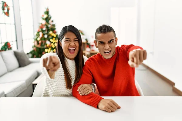 Young Latin Couple Sitting Table Christmas Tree Pointing Displeased Frustrated — Stockfoto