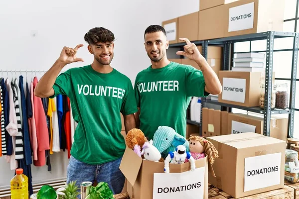 Young gay couple wearing volunteer t shirt at donations stand smiling and confident gesturing with hand doing small size sign with fingers looking and the camera. measure concept.
