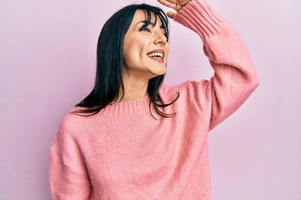 Young brunette woman with bangs wearing casual winter sweater very happy and smiling looking far away with hand over head. searching concept.
