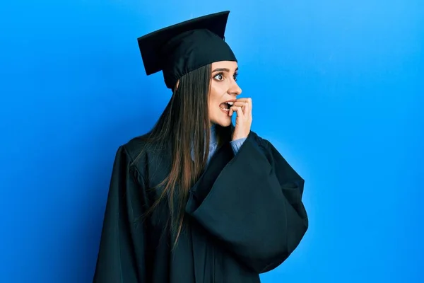 卒業キャップと式のローブを身に着けている美しいブルネットの若い女性は 口の中に爪をかむ上で手で強調し 緊張している 不安の問題 — ストック写真