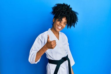 Afro saçlı, karate kimonosu ve siyah kuşak giyen Afro-Amerikalı bir kadın elleriyle mutlu bir jest yapıyor. Kameraya bakıp başarıyı gösteren ifade. 