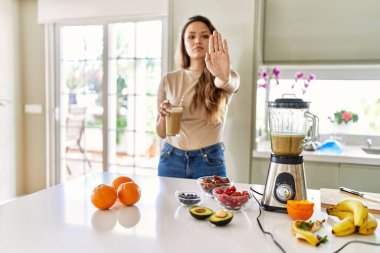 Güzel, genç, esmer bir kadın mutfakta açık elle bir bardak smoothie içiyor dur işareti yapıyor ciddi ve kendinden emin bir ifadeyle, savunma hareketi yapıyor. 