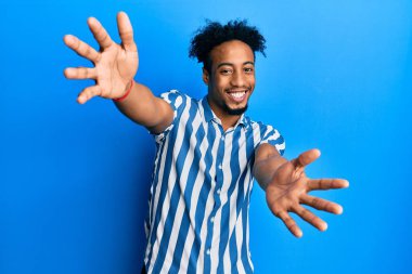 Sakallı, sıradan çizgili tişörtlü genç Afro-Amerikan adam kameraya bakıp sarılmak için gülümsüyor. Mutluluğu kucaklayan neşeli bir ifade.. 