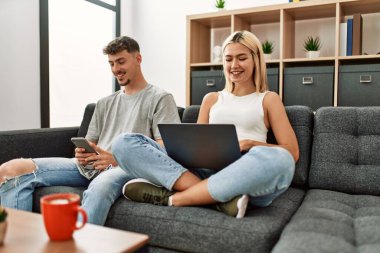 Evdeki kanepede oturan genç beyaz çift dizüstü bilgisayar ve akıllı telefon kullanarak mutlu bir şekilde gülümsüyor..