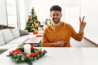 Arap genç adam, Noel ağacının yanında oturmuş kameraya gülümseyen ve zafer işareti yapan gülümsüyor. İki numara.. 