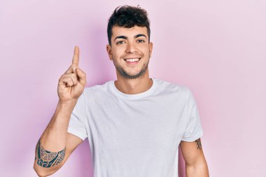 Günlük beyaz tişört giyen genç İspanyol bir adam bir fikir ya da soruyla gülümsüyor ve mutlu bir yüzle işaret ediyor. 