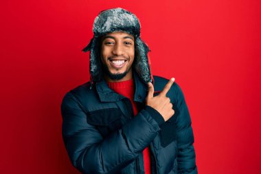 Sakallı, kışlık şapkalı ve ceketli, yüzünde mutlu ve doğal bir ifadeyle elleri ve parmaklarıyla yüzünü gösteren gülümseyen Afrikalı genç bir Amerikalı. 