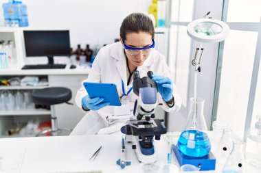 Laboratuvarda dokunmatik ped ve mikroskop kullanarak bilim adamı üniforması giyen İspanyol genç bir kadın.