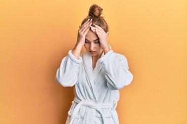 Bornoz giyen genç sarışın kadın baş ağrısı çekiyor. Acı ve migren yüzünden çaresiz ve stresli. Eller başının üstünde.. 