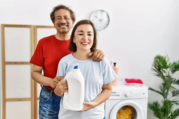 中年跨种族夫妇一边洗衣服一边拿着洗涤剂瓶子 看起来很积极 站着很快乐 微笑着 带着自信的笑容露出牙齿 — 图库照片