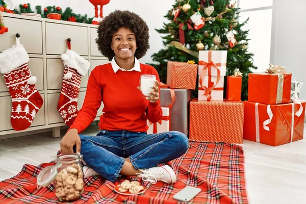若いですアフリカ系アメリカ人女性とともにアフロヘア食べるペストリーによって座ってクリスマスツリー見て正と幸せな立ちと笑顔で自信を持って笑顔示す歯 — ストック写真