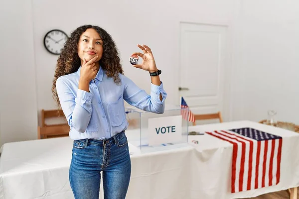 Beautiful hispanic woman standing by at political campaign by voting ballot looking confident at the camera smiling with crossed arms and hand raised on chin. thinking positive.