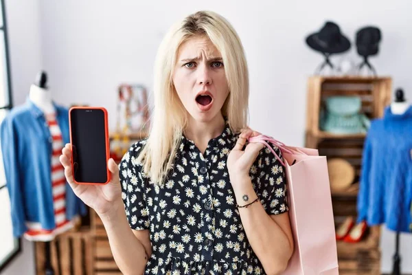 若いですブロンド女性を保持ショッピングバッグを示すスマートフォンの画面恐怖と驚きと驚きの表情でショックを受けました 恐怖と興奮した顔 — ストック写真