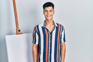 Yüzünde mutlu ve serin bir gülümsemeyle boş tuvalin yanında duran genç bir İspanyol. şanslı kişi. 