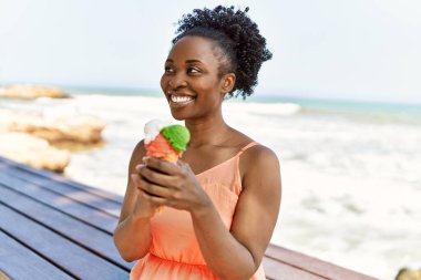 Genç Afrikalı Amerikalı bir kadın kumsalda mutlu bir şekilde gülümsüyor ve dondurma yiyor.