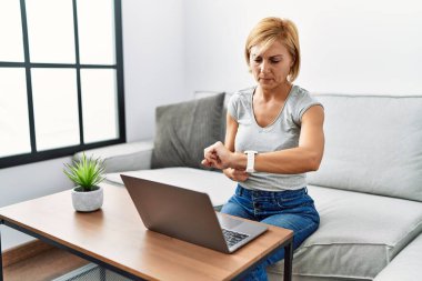 Orta yaşlı sarışın kadın evde dizüstü bilgisayar kullanıyor. Saati kontrol ediyor, rahat ve kendinden emin. 