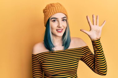 Genç modern kız yün şapka takıyor ve parmaklarıyla beş numarayı gösterirken kendinden emin ve mutlu gülümsüyor.. 