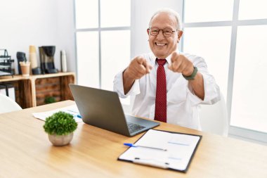 Ofiste çalışan kıdemli bir adam bilgisayarı kullanarak seni ve kamerayı işaret ediyor ve gülümsüyor. 