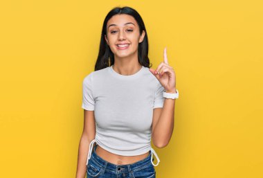 Gündelik beyaz tişört giyen genç İspanyol kız bir numaralı parmağını gösterip işaret ederken kendine güvenen ve mutlu bir şekilde gülümsüyor.. 