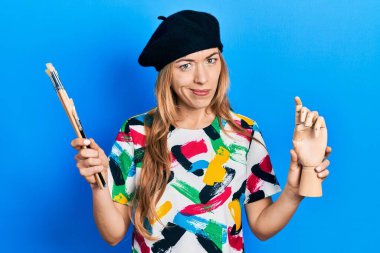 Kafkasyalı genç bir kadın sanatçı elinde küçük ahşap bir manikin ve boya fırçaları tutuyor şüpheci ve gergin, sorun yüzünden surat asıyor. negatif kişi. 