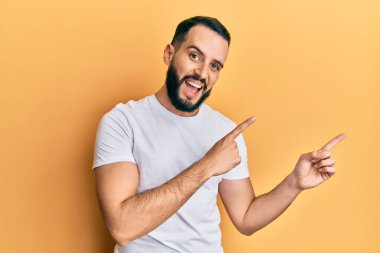 Sakallı, günlük beyaz tişörtlü, gülümseyen ve kameraya bakan genç bir adam.. 