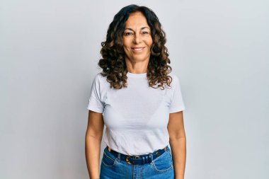 Orta yaşlı İspanyol kadın, mutlu ve havalı bir gülümsemesi olan günlük beyaz tişört giyiyor. şanslı kişi. 