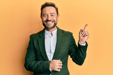 Orta yaşlı takım elbiseli, yüzünde kocaman bir gülümseme olan, eli ve parmağı kameraya bakan bir adam.. 