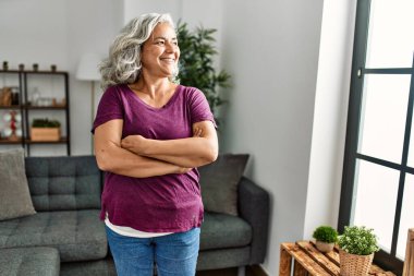 Orta yaşlı, gri saçlı, gülümseyen, mutlu, kollarını kavuşturmuş bir kadın..