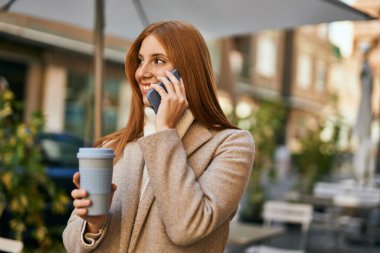 Genç kızıl saçlı kız akıllı telefonda konuşuyor. Şehirde kahve içiyor..