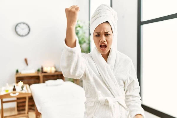 Young brunette woman wearing towel and bathrobe standing at beauty center angry and mad raising fist frustrated and furious while shouting with anger. rage and aggressive concept.