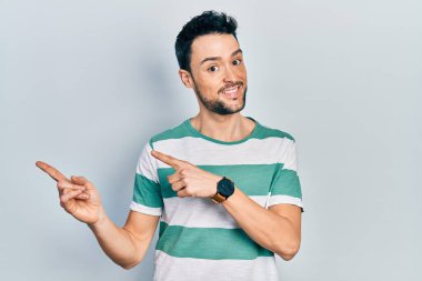 Gündelik kıyafetler giyen genç İspanyol adam gülümseyip kameraya bakıyor ve iki eliyle ve parmaklarıyla kenarı işaret ediyor.. 